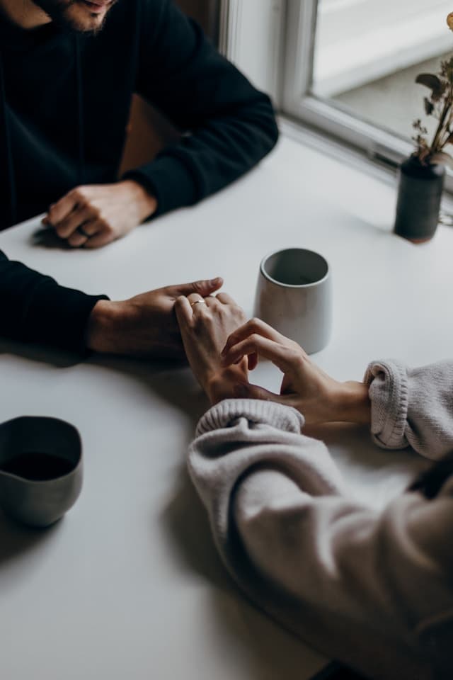 Two people sitting at a table, gently holding hands in an intimate moment. Mugs of coffee are on the table, and soft natural light streams in from a nearby window, creating a peaceful and tender atmosphere.