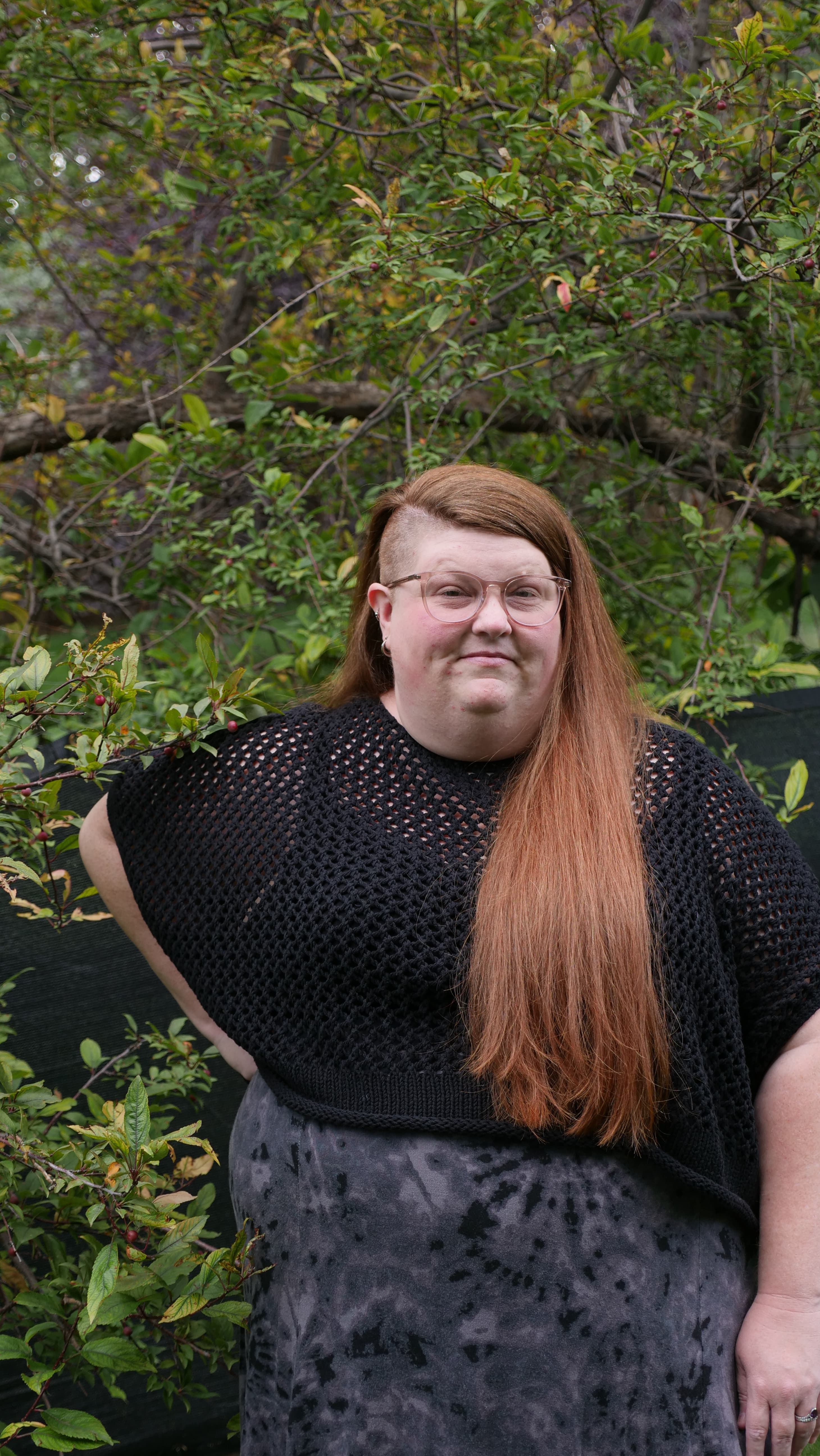 Jamie L. Thompson stands confidently in front of lush green foliage. She has long, reddish-brown hair and glasses, wearing a black, textured top, and offering a calm, relaxed expression.